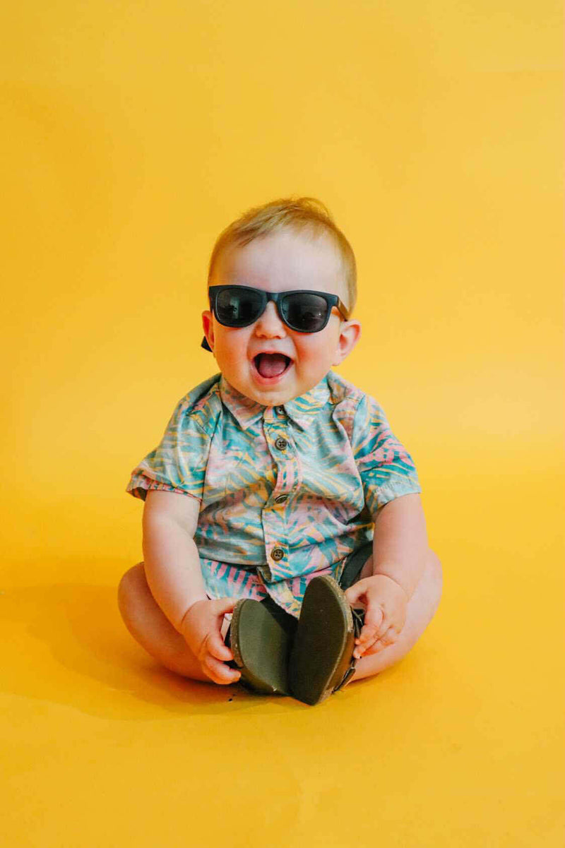 Toddler wearing black sunglasses while sitting against a yellow background.
